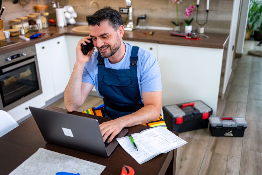 A home service business owner on the phone at a kitchen table, laptop open, tools beside him